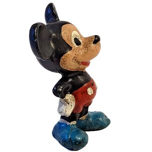 May include: A vintage-style molded figurine of a cartoon mouse character with large black ears, a tan face, red shorts with white buttons, and blue shoes. The paint shows signs of wear and chipping, revealing the texture of the material underneath.