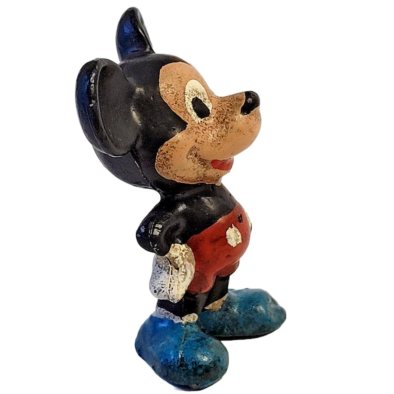 May include: A vintage-style molded figurine of a cartoon mouse character with large black ears, a tan face, red shorts with white buttons, and blue shoes. The paint shows signs of wear and chipping, revealing the texture of the material underneath.
