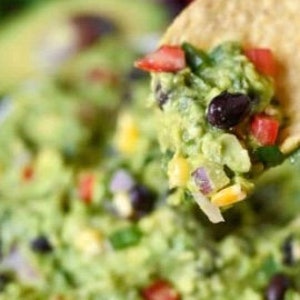 May include: Close-up of guacamole with a tortilla chip scooping up a portion. The guacamole is a vibrant green color and contains visible pieces of red tomatoes, black beans, and yellow corn. The chip is golden brown.
