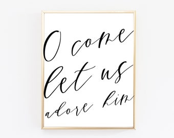 May include: A framed print with the words "O come let us adore him" in elegant black script. The text is centered on a white background, framed by a thin gold border, and set against a plain white backdrop.