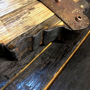 May include: Close-up of a wooden board with a metal strap and copper rivets. The wood is dark brown and has a rough texture. The metal strap is rusty and has a worn look.