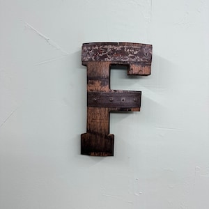 Puede incluir: Una letra F de madera hecha de madera recuperada con detalles metálicos. La letra es de color marrón oscuro y tiene un aspecto rústico.