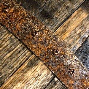 May include: A close-up of a rustic wooden surface with a rusty metal band. The band is secured with several copper screws. The wood has a natural grain pattern and is stained a dark brown color.