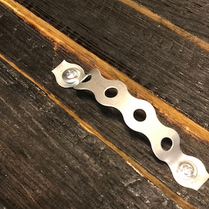 May include: A silver metal hanging bracket with a wavy design and screw holes. The bracket is attached to a dark brown wooden surface.