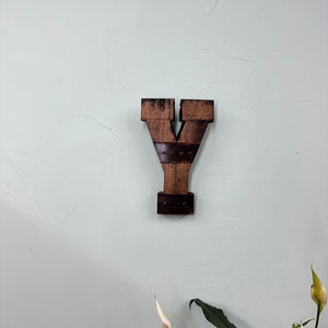 May include: A wooden letter Y made from reclaimed wood, mounted on a light green wall. The letter is dark brown and has a rustic appearance.