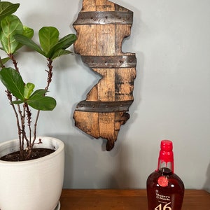 May include: A wooden state of New Jersey outline, made from reclaimed bourbon barrel staves, with metal bands and rivets. The state outline is mounted on a wall, with a bottle of Maker's Mark 46 bourbon whiskey in the foreground.
