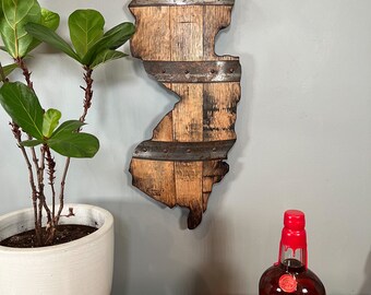 May include: A wooden state of New Jersey outline, made from reclaimed bourbon barrel staves, with metal bands and rivets. The state outline is mounted on a wall, with a bottle of Maker's Mark 46 bourbon whiskey in the foreground.