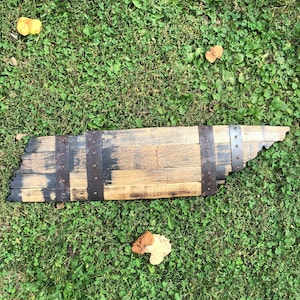 May include: A wooden sign shaped like the state of Tennessee, made from a reclaimed whiskey barrel stave. The sign is stained dark brown and has metal bands around the edges.