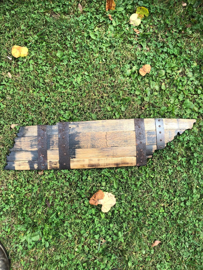 May include: A wooden sign shaped like the state of Tennessee, made from a reclaimed whiskey barrel stave. The sign is stained dark brown and has metal bands around the edges.