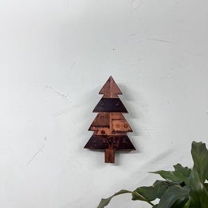 May include: A wooden Christmas tree decoration with a rustic aesthetic. The tree is made of various wood pieces in shades of brown and dark brown, with a triangular shape. It is mounted on a white wall, with a green plant in the bottom right corner.