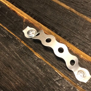 May include: A silver metal hanging bracket with a wavy design and screw holes. The bracket is attached to a dark brown wooden surface.