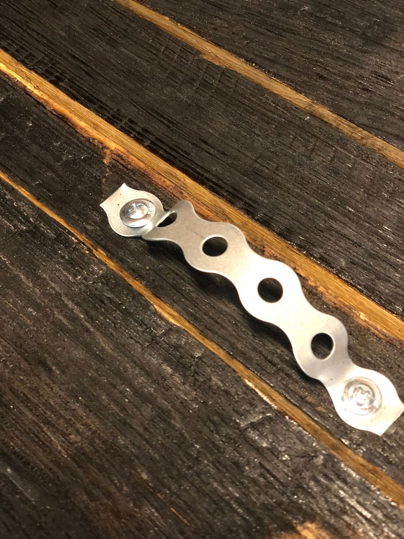 May include: A silver metal hanging bracket with a wavy design and screw holes. The bracket is attached to a dark brown wooden surface.