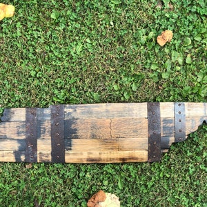 May include: A wooden state of Tennessee outline, made from reclaimed bourbon barrel staves. The wood is a mix of brown and black, with metal bands and rivets.