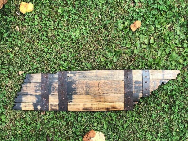 May include: A wooden state of Tennessee outline, made from reclaimed bourbon barrel staves. The wood is a mix of brown and black, with metal bands and rivets.