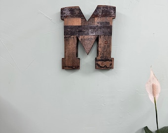 May include: A wooden letter M, crafted from reclaimed wood, with a dark brown stain and metal accents. The letter is mounted on a light green wall.
