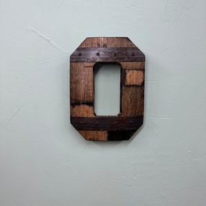 May include: A wooden letter O, crafted from reclaimed wood and accented with metal bands. The letter is painted a dark brown color and has a rustic, industrial look.