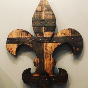 May include: A wooden fleur-de-lis wall decor made from reclaimed wood with copper accents. The wood has a dark brown stain and is distressed.