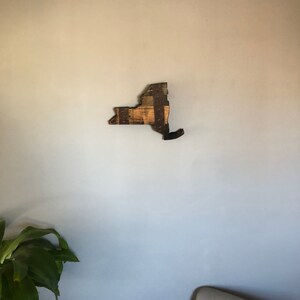 May include: A wooden wall art piece in the shape of the state of New York. The wood is a light brown color and has a distressed look. The piece is mounted on a light blue wall.