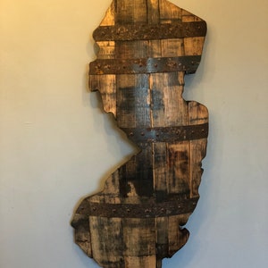 May include: Wooden wall art shaped like the state of New Jersey. The artwork is constructed from reclaimed wood planks and held together with metal bands. The wood has a weathered, rustic appearance with varying shades of brown and gray.