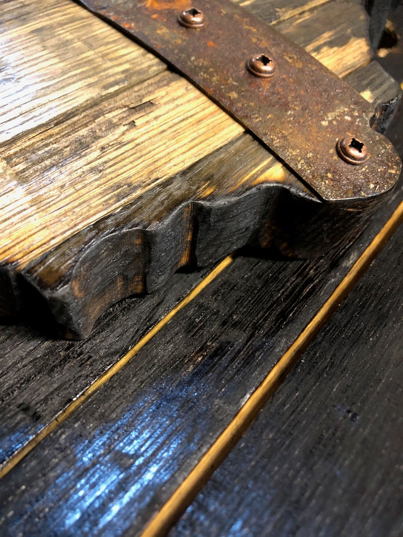 May include: Close-up of a wooden board with a metal strap and copper rivets. The wood is dark brown and has a rough texture. The metal strap is rusty and has a worn look.