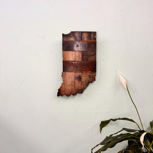 May include: A wooden wall art piece shaped like the state of Indiana. The wood is a dark brown color and has a rustic look. The piece is decorated with metal bands and rivets.
