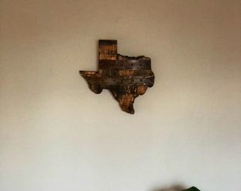 May include: A wooden wall art piece in the shape of the state of Texas. The wood is dark brown and has a rustic look. The piece is mounted on a white wall.