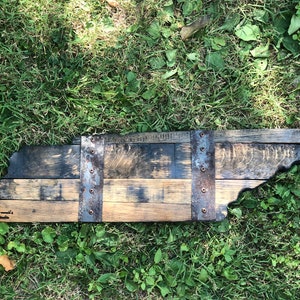 May include: A wooden state of Tennessee outline, made from reclaimed bourbon barrel staves. The state is outlined in dark wood and has metal bands with rivets. The words "Tennessee Made" are written on the bottom left corner of the state.