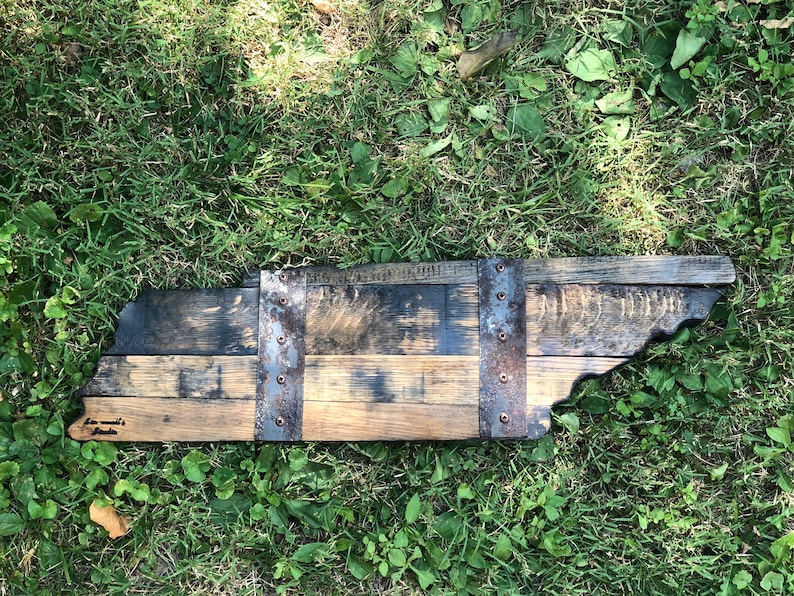 May include: A wooden state of Tennessee outline, made from reclaimed bourbon barrel staves. The state is outlined in dark wood and has metal bands with rivets. The words "Tennessee Made" are written on the bottom left corner of the state.