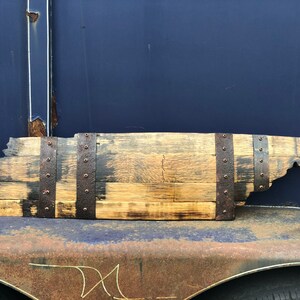 May include: A wooden state of Tennessee outline, crafted from a reclaimed whiskey barrel stave. The wood is stained a dark brown and has a rustic, weathered look. The state outline is secured with metal bands and rivets.