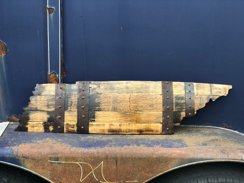 May include: A wooden state of Tennessee outline, crafted from a reclaimed whiskey barrel stave. The wood is stained a dark brown and has a rustic, weathered look. The state outline is secured with metal bands and rivets.