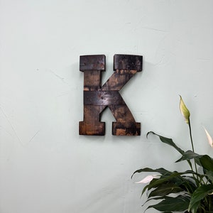 May include: A dark brown wooden letter K, made from reclaimed wood, with metal accents. The letter is mounted on a light green wall.