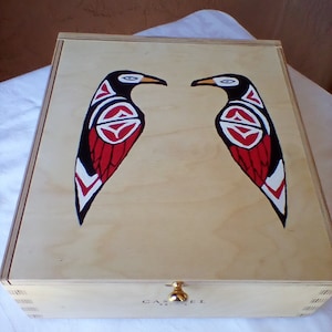 May include: Wooden box with a light wood finish, featuring two stylized bird designs in black, red, and white. The birds face each other. The box has a gold-colored clasp and the word "CASTEL" is printed on the front.
