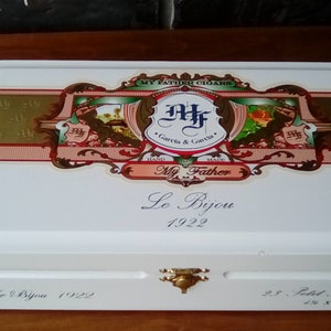 May include: White wooden box with a gold-trimmed lid. The lid features a colorful label with the text "My Father Cigars", "Garcia & Garcia", "My Father", and "Le Bijou 1922". The bottom of the box has the text "Le Bijou 1922" and "25. Petit Robusto 4.8 x 52".