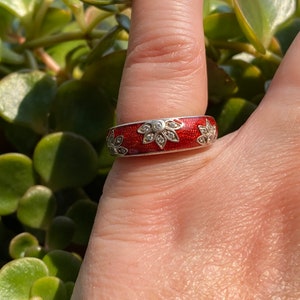 May include: A silver ring with a red enamel band. The ring features a floral design with clear gemstones. The ring is shown on a finger with a green leafy background.