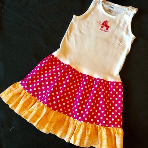 May include: A sleeveless dress with a white bodice featuring a small embroidered red roller skate and the text let's roll. The skirt is bright pink with white polka dots and a ruffled hem in a yellow fabric with a white floral pattern.