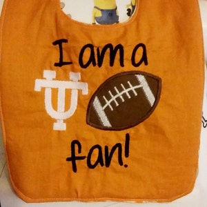 May include: Orange fabric baby bib with the text "I am a " followed by a white University of Tennessee logo and a brown football with white laces. The text "fan!" is below the logo and football.