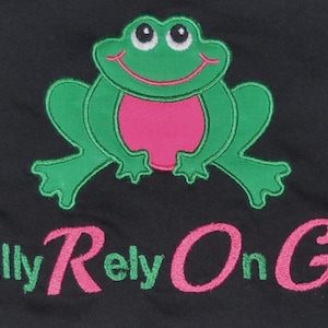 Może przedstawiać: Czarny T-shirt z haftowanym zielonym żabą z przodu. Żaba ma różowe akcenty i szeroki uśmiech. Tekst "Fully Rely On God" jest wyszyty pod żabą na różowo i zielono.