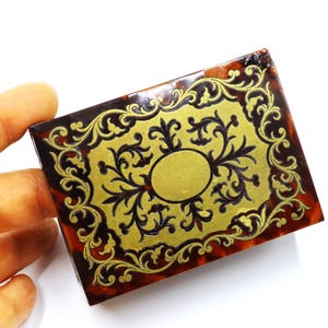 May include: A small, rectangular box made of tortoiseshell with a gold-colored design. The design features a central oval shape surrounded by intricate floral patterns.
