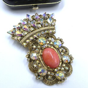 May include: Vintage gold-tone brooch with a crown design, featuring iridescent rhinestones and a central oval coral-colored stone. The brooch has an ornate, filigree-style setting and is likely from the mid-20th century. The brooch is a statement piece.