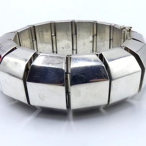 Puede incluir: Un brazalete de plata con un diseño geométrico. El brazalete está hecho de eslabones entrelazados que forman una serie de formas redondeadas y angulares.