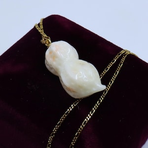 May include: A white peanut-shaped pendant necklace with a gold chain. The pendant has a slightly pink hue and is made of a smooth, polished material.