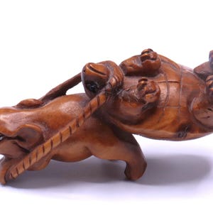 May include: A detailed wooden carving of a frog pulling a turtle on its back. The frog is brown with dark eyes and a rope attached to the turtle. The turtle is on its back, with its legs in the air. The carving is set against a white background.