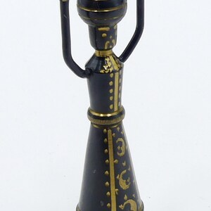 May include: A small, black and gold metal figurine of a person with raised arms. The figurine is wearing a long, black robe with gold accents and a gold crown. The figurine is standing on a small, round base. The figurine has a tag attached to the base that says "Origin Lebanon".