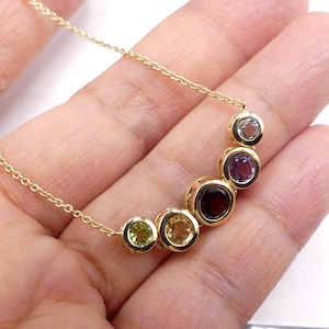 May include: A gold chain necklace with a rainbow of gemstones. The stones are set in gold and are arranged in a crescent shape. The stones are green, yellow, orange, red, purple, and clear.