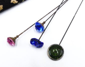 May include: Four vintage hat pins with decorative glass heads. The heads are blue, pink, green, and a darker blue. The pins are made of metal and have a rusty appearance.
