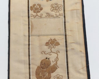 May include: A beige silk embroidered wall hanging with a black border. The hanging features two figures in brown, each holding a flower. The figures are surrounded by floral designs.