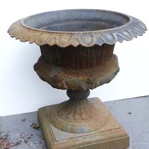 May include: A large, antique, cast iron planter with a square base and a fluted, urn-shaped body. The planter is weathered and shows signs of rust.