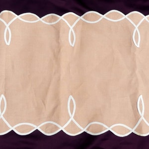 May include: A beige table runner with white embroidered floral designs and scalloped edges.