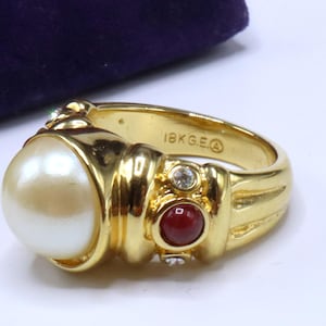 May include: A gold ring with a large cream-coloured pearl and a smaller, round, red stone. The ring also has two clear, round accent stones and is marked "18K G.E."