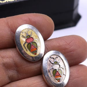 May include: Oval silver cufflinks with a colorful insect design. The insects have red, green, and orange details on a cream-colored background. The cufflinks are set against a white background.